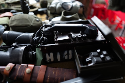 A collection of vintage military items including a field radio with a rotary dial, a pair of binoculars, and a knife with a leather sheath. The objects are arranged on a surface, with some additional gear partially visible in the background.