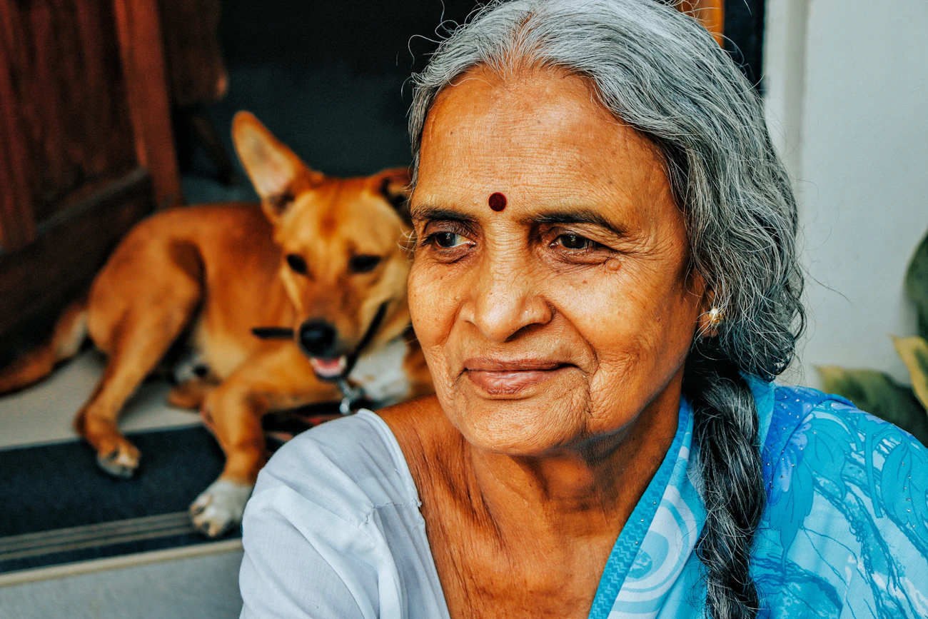 A calm older woman with gray hair sitting in a doorway beside a small tan dog resting at her side