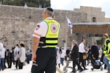 A paramedic wearing a high-visibility vest stands among a group of people at a historical or religious site. The background includes an ancient stone wall and a portion of a covered area, with an Israeli flag visible. Several people in traditional clothing, possibly engaged in prayer or conversation, can be seen.