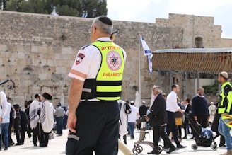A paramedic wearing a high-visibility vest stands among a group of people at a historical or religious site. The background includes an ancient stone wall and a portion of a covered area, with an Israeli flag visible. Several people in traditional clothing, possibly engaged in prayer or conversation, can be seen.