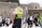 A paramedic wearing a high-visibility vest stands among a group of people at a historical or religious site. The background includes an ancient stone wall and a portion of a covered area, with an Israeli flag visible. Several people in traditional clothing, possibly engaged in prayer or conversation, can be seen.