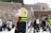 A paramedic wearing a high-visibility vest stands among a group of people at a historical or religious site. The background includes an ancient stone wall and a portion of a covered area, with an Israeli flag visible. Several people in traditional clothing, possibly engaged in prayer or conversation, can be seen.