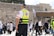 A paramedic wearing a high-visibility vest stands among a group of people at a historical or religious site. The background includes an ancient stone wall and a portion of a covered area, with an Israeli flag visible. Several people in traditional clothing, possibly engaged in prayer or conversation, can be seen.