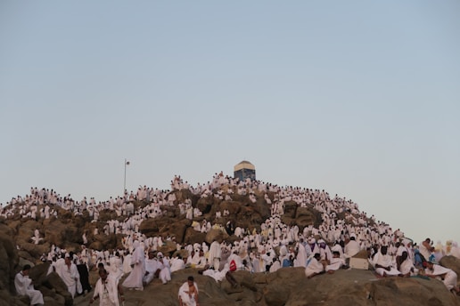 A large crowd of people wearing white garments gather closely together on a rocky hill under a clear sky. The scene is one of congregation and possibly a religious or spiritual event, with most individuals seated or standing among the rocks.
