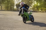 A motorcyclist performing a stunt on a green sports bike, balancing on the rear wheel. The rider is wearing protective gear, including a red and white helmet, and is in an open area surrounded by trees.