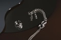 A collection of silver jewelry, including a chunky ring and a thick chain bracelet, is arranged on a glossy, dark surface. The reflective surface creates shimmering highlights and enhances the metallic sheen of the pieces.