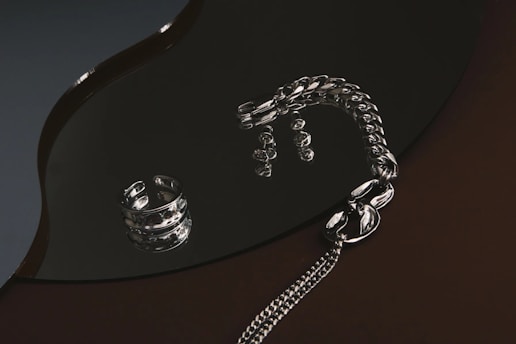 A collection of silver jewelry, including a chunky ring and a thick chain bracelet, is arranged on a glossy, dark surface. The reflective surface creates shimmering highlights and enhances the metallic sheen of the pieces.
