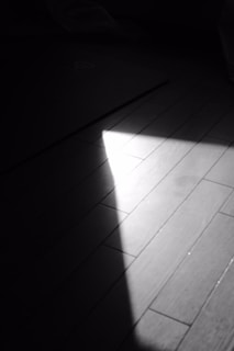 A beam of light illuminates a portion of a wooden floor, creating a striking contrast between light and shadow. The floorboards are visible with subtle textures and lines, adding depth to the image.