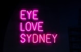 Bright pink neon letters spell out the phrase 'EYE LOVE SYDNEY' against a dark background, creating a bold and attractive contrast.