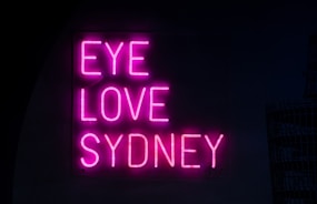 Bright pink neon letters spell out the phrase 'EYE LOVE SYDNEY' against a dark background, creating a bold and attractive contrast.