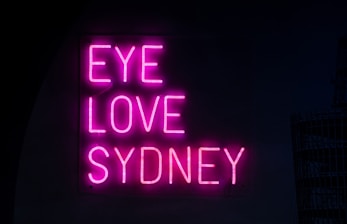 Bright pink neon letters spell out the phrase 'EYE LOVE SYDNEY' against a dark background, creating a bold and attractive contrast.