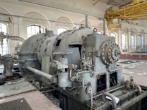 An industrial setting features a large, old machine with various pipes, valves, and rusted metal components. The room has high arched windows allowing natural light, exposed pipes on the walls, and a yellow overhead crane system. The floor is littered with debris, indicating the space is unused or abandoned.