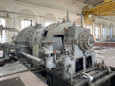 An industrial setting features a large, old machine with various pipes, valves, and rusted metal components. The room has high arched windows allowing natural light, exposed pipes on the walls, and a yellow overhead crane system. The floor is littered with debris, indicating the space is unused or abandoned.