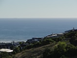 A serene coastal scene with houses nestled on a hillside, overlooking a calm ocean that extends to the horizon. The landscape is lush with green vegetation, and the clear sky suggests a peaceful day.