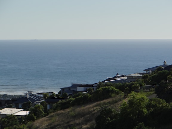 A serene coastal scene with houses nestled on a hillside, overlooking a calm ocean that extends to the horizon. The landscape is lush with green vegetation, and the clear sky suggests a peaceful day.