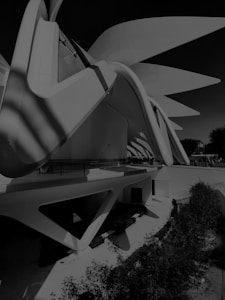 A modern architectural structure with a dynamic, futuristic design. The building features large, sweeping curves and sharp angles, creating an impression of movement and innovation. Shadows and light play across the surfaces, adding depth and contrast. The surrounding environment includes lush greenery, enhancing the juxtaposition of nature with the architectural elements.
