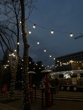 A street food truck with the sign 'STREET CHEFS' is set up in an outdoor area during the evening. Fairy lights are strung across tree branches, creating a warm and inviting ambiance. There are outdoor heaters and high tables around, with a few people gathered near the truck.