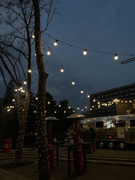 A street food truck with the sign 'STREET CHEFS' is set up in an outdoor area during the evening. Fairy lights are strung across tree branches, creating a warm and inviting ambiance. There are outdoor heaters and high tables around, with a few people gathered near the truck.