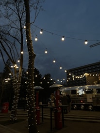A street food truck with the sign 'STREET CHEFS' is set up in an outdoor area during the evening. Fairy lights are strung across tree branches, creating a warm and inviting ambiance. There are outdoor heaters and high tables around, with a few people gathered near the truck.
