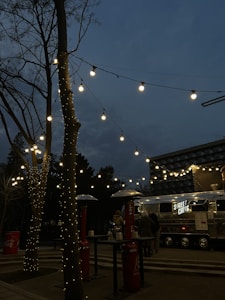 A street food truck with the sign 'STREET CHEFS' is set up in an outdoor area during the evening. Fairy lights are strung across tree branches, creating a warm and inviting ambiance. There are outdoor heaters and high tables around, with a few people gathered near the truck.