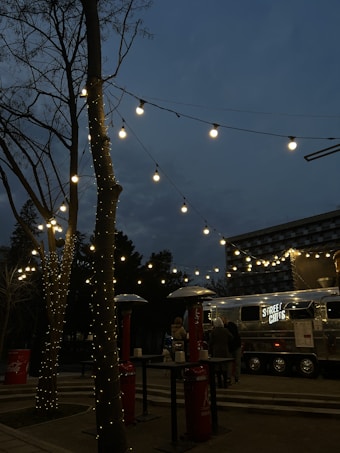 A street food truck with the sign 'STREET CHEFS' is set up in an outdoor area during the evening. Fairy lights are strung across tree branches, creating a warm and inviting ambiance. There are outdoor heaters and high tables around, with a few people gathered near the truck.
