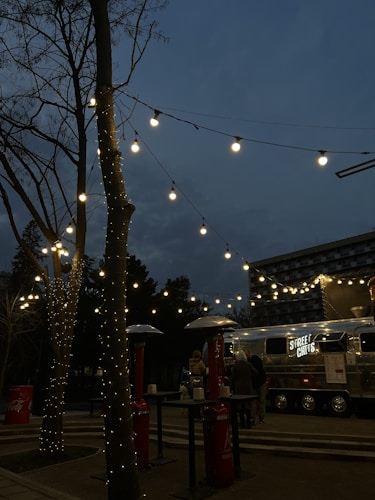 A street food truck with the sign 'STREET CHEFS' is set up in an outdoor area during the evening. Fairy lights are strung across tree branches, creating a warm and inviting ambiance. There are outdoor heaters and high tables around, with a few people gathered near the truck.