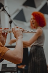 A person is playing a flute, focusing on their hands and the instrument. In the background, another person with bright red curly hair is slightly out of focus, possibly singing or speaking. The setting appears to be indoors with some geometric decorations or panels on the wall.