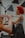 A person is playing a flute, focusing on their hands and the instrument. In the background, another person with bright red curly hair is slightly out of focus, possibly singing or speaking. The setting appears to be indoors with some geometric decorations or panels on the wall.