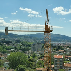 A large yellow construction crane is prominently featured in front of a cityscape. The background includes a mix of urban and natural landscapes with buildings, houses, and green trees. Rolling hills and mountains can be seen in the distance under a blue sky with scattered clouds.