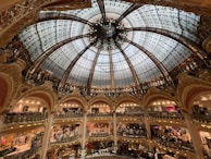 A grand, intricately designed glass dome ceiling with ornate detailing hovers over multiple levels of a luxury shopping mall. The architecture is elaborate, with decorative arches and railings. The space is bustling with people shopping across various floors.