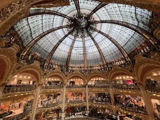 A grand, intricately designed glass dome ceiling with ornate detailing hovers over multiple levels of a luxury shopping mall. The architecture is elaborate, with decorative arches and railings. The space is bustling with people shopping across various floors.