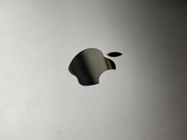 A metallic surface with a prominent apple-shaped logo etched into it. The logo is sleek and reflective, set against a smooth, matte background.