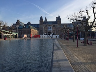A large building with traditional European architectural features stands prominently in the background. In front, a reflecting pool stretches along a paved walkway. Leafless trees line the sides, and a few people are walking nearby. The famous 'I amsterdam' sign is placed before the building.