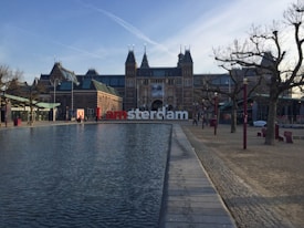 A large building with traditional European architectural features stands prominently in the background. In front, a reflecting pool stretches along a paved walkway. Leafless trees line the sides, and a few people are walking nearby. The famous 'I amsterdam' sign is placed before the building.