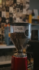 A coffee grinder filled with coffee beans is prominently displayed, featuring a transparent hopper with a logo for Wise Coffee. The background is blurred, showcasing a collage of black-and-white images and some colorful elements.