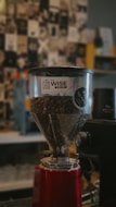 A coffee grinder filled with coffee beans is prominently displayed, featuring a transparent hopper with a logo for Wise Coffee. The background is blurred, showcasing a collage of black-and-white images and some colorful elements.