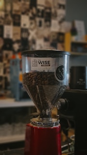 A coffee grinder filled with coffee beans is prominently displayed, featuring a transparent hopper with a logo for Wise Coffee. The background is blurred, showcasing a collage of black-and-white images and some colorful elements.
