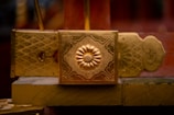A close-up of an ornate golden decoration featuring an embossed flower design in the center, surrounded by intricate patterns. The background shows additional gold and red elements with similar patterns.