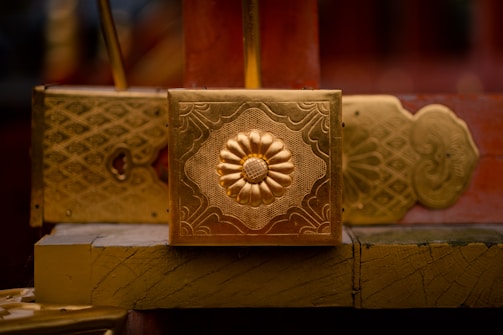 A close-up of an ornate golden decoration featuring an embossed flower design in the center, surrounded by intricate patterns. The background shows additional gold and red elements with similar patterns.