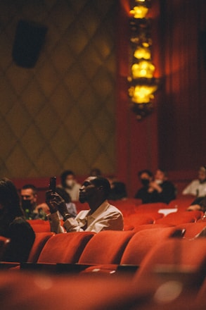 A dimly lit theater with red seats and ornate wall lights. The focus is on a person in the foreground holding a smartphone, dressed in a light-colored shirt. Other people are seated in the background, some wearing face masks.