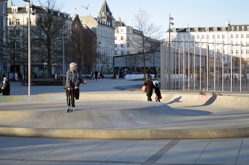A skate park in an urban setting, surrounded by white buildings and leafless trees. There are several people in the area, including a person on a scooter and two others walking nearby. The sun casts long shadows on the ground, indicating late afternoon or early evening. A bird is flying in the sky.