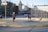 A skate park in an urban setting, surrounded by white buildings and leafless trees. There are several people in the area, including a person on a scooter and two others walking nearby. The sun casts long shadows on the ground, indicating late afternoon or early evening. A bird is flying in the sky.