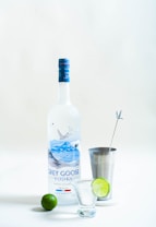 A bottle of Grey Goose vodka stands upright next to a metal cocktail shaker with a stirring stick and a lime slice. In front of the vodka bottle, there is a small glass and a whole lime. The scene is set against a plain, light background.
