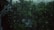Raindrops on a glass window with a view of a lush green forest or garden. The scene appears dark and moody, likely due to overcast weather. An eavestrough is visible in the top left corner.