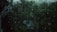 Raindrops on a glass window with a view of a lush green forest or garden. The scene appears dark and moody, likely due to overcast weather. An eavestrough is visible in the top left corner.