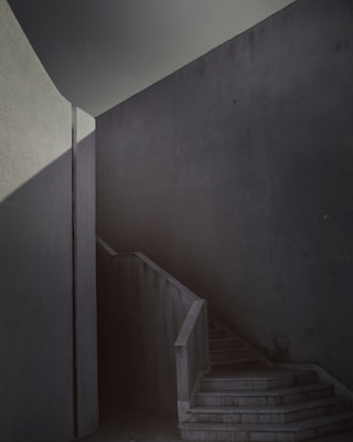 A minimalistic interior staircase with sharp geometric lines and shadows. The space is enclosed by high, plain walls with a subdued texture, and the lighting creates a dramatic play of light and shade.
