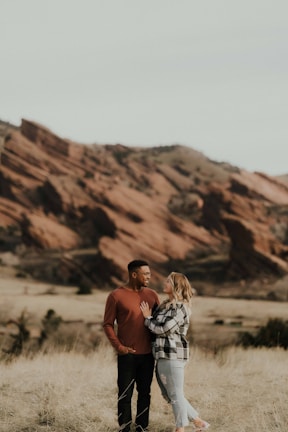 A couple stands together in a grassy field with rocky formations in the background. The man is wearing a red shirt and the woman is in a black-and-white checked shirt with jeans. They are looking into each other's eyes, conveying a sense of closeness.