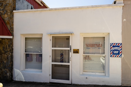 A small, white-painted building with two rectangular windows on either side of a glass door. The sign above the door reads 'Leatherman's Barber Shop Since 1915'. There are American flags hanging inside the left window and a sign on the right window indicates the shop's hours. A barber pole can be seen behind the glass door. Another sign with a barber's pole design is mounted on the wall to the right of the door.
