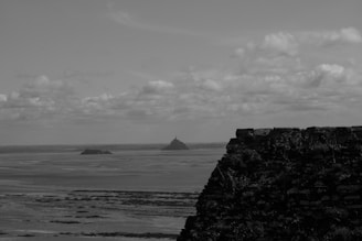 A black and white landscape photograph featuring a coastal scene with distant islands and a prominent rock structure in the foreground. The sky is partly cloudy, casting shadows over the flat expanse of water and shore.