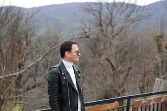 Model wearing a sleek black leather jacket standing against a rugged mountain backdrop.